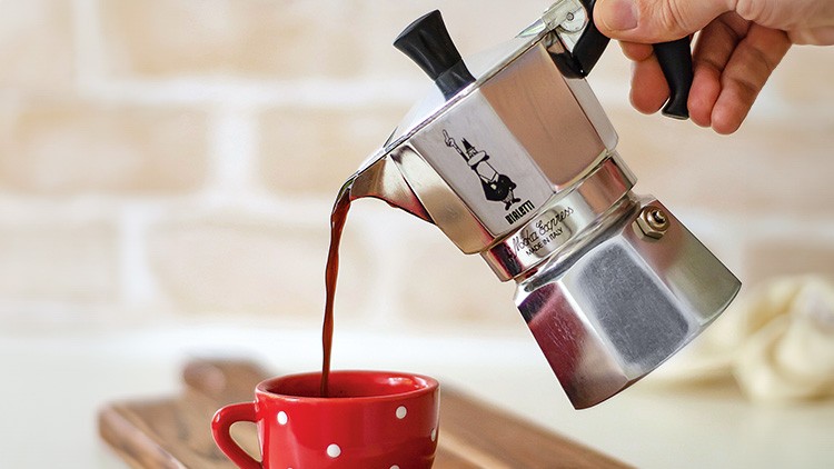 Eine silberne Espressokanne gießt Kaffee in eine rote Tasse mit weißen Punkten, Holzbrett und helle Wand im Hintergrund.