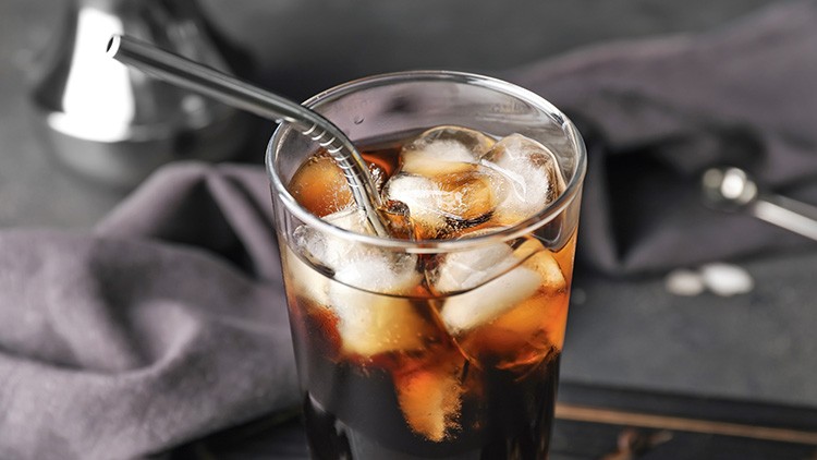 Ein Glas mit Cola, Eiswürfeln und einem Metalltrinkhalm steht vor einem dunklen Hintergrund mit Stoff und unscharfem Zubehör.