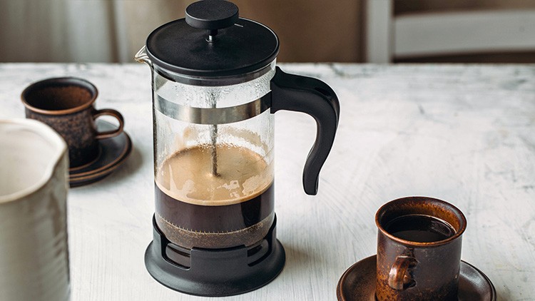 Eine schwarze Pressstempelkanne mit Kaffee steht auf einem weißen Tisch, daneben zwei braune Tassen und ein weißer Krug.