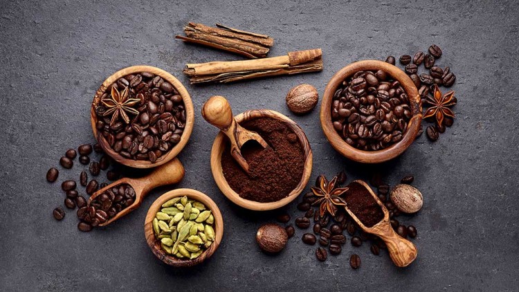 Verschiedene Holzgefäße mit Kaffeebohnen, gemahlenem Kaffee, Kardamom, Zimtstangen, Sternanis und Muskatnüssen auf dunklem Untergrund.