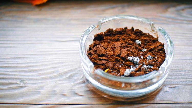 Durchsichtiger Aschenbecher mit dunkelbraunem Kaffeepulver und silbrig glänzenden Resten auf einer Holzoberfläche.