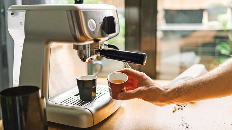 Silberne Espressomaschine auf Holztisch bereitet zwei Espressi zu, während eine Hand eine kleine rote Tasse hält.