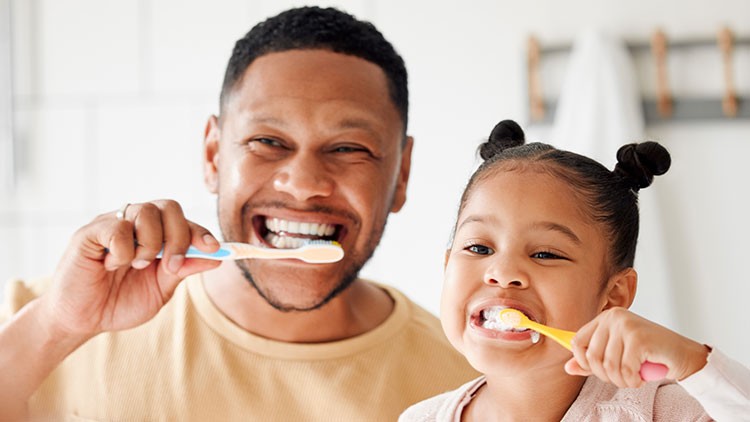 Zwei Personen putzen sich die Zähne nebeneinander in einem hellen Badezimmer, im Hintergrund hängen Handtücher.