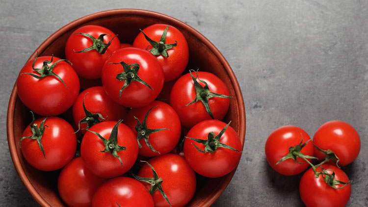 Rote Tomaten mit grünen Stielen liegen in einer braunen Schale auf einem grauen Untergrund.