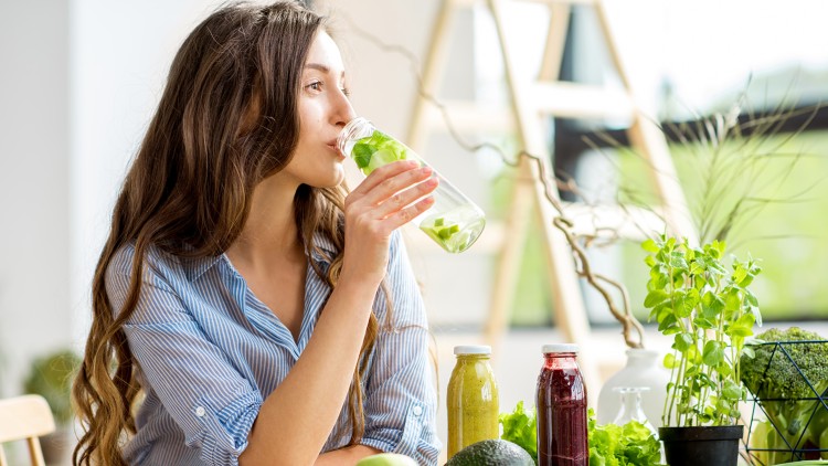 Person hält eine Flasche mit Wasser und Limette, auf einem Tisch liegen frisches Obst, Gemüse, Nüsse und Smoothies.